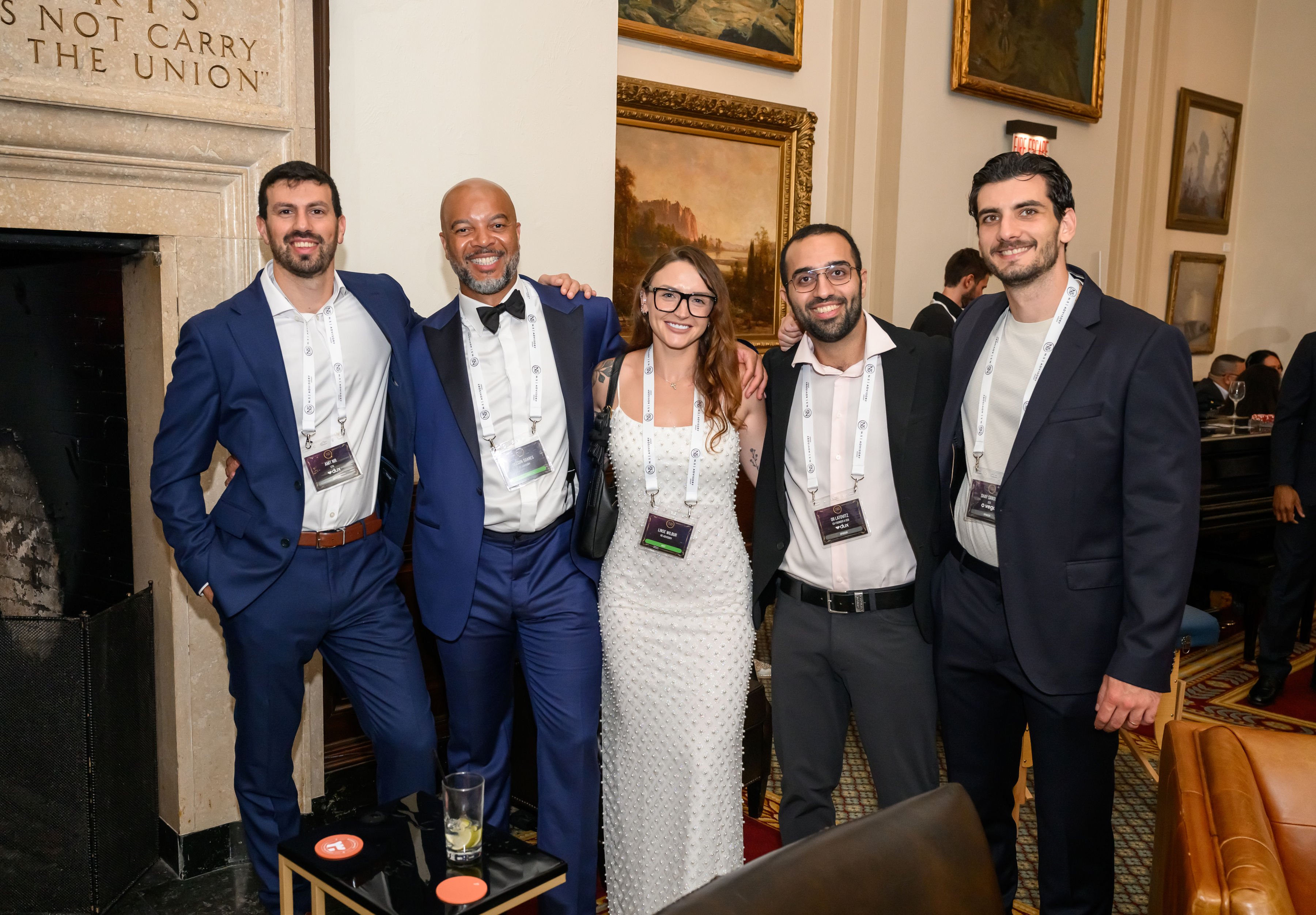 Five guests in formal attire smiling together at an Astra evening, wearing lanyards beside a fireplace in an oak-panelled room hung with gilt-framed paintings.
