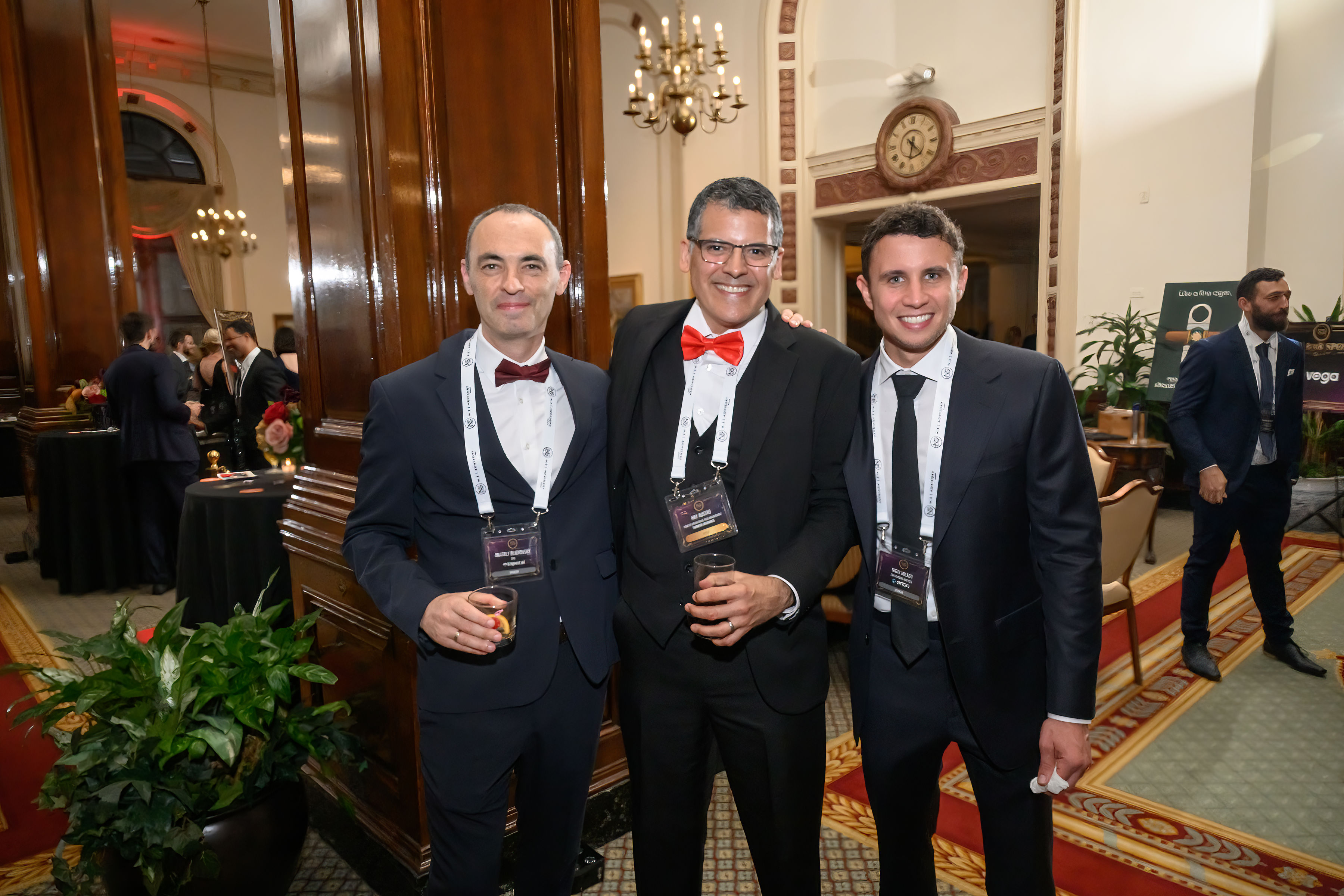 Three executives in black-tie attire smiling together at an Astra evening, wearing event lanyards in an oak-panelled room lit by a chandelier.