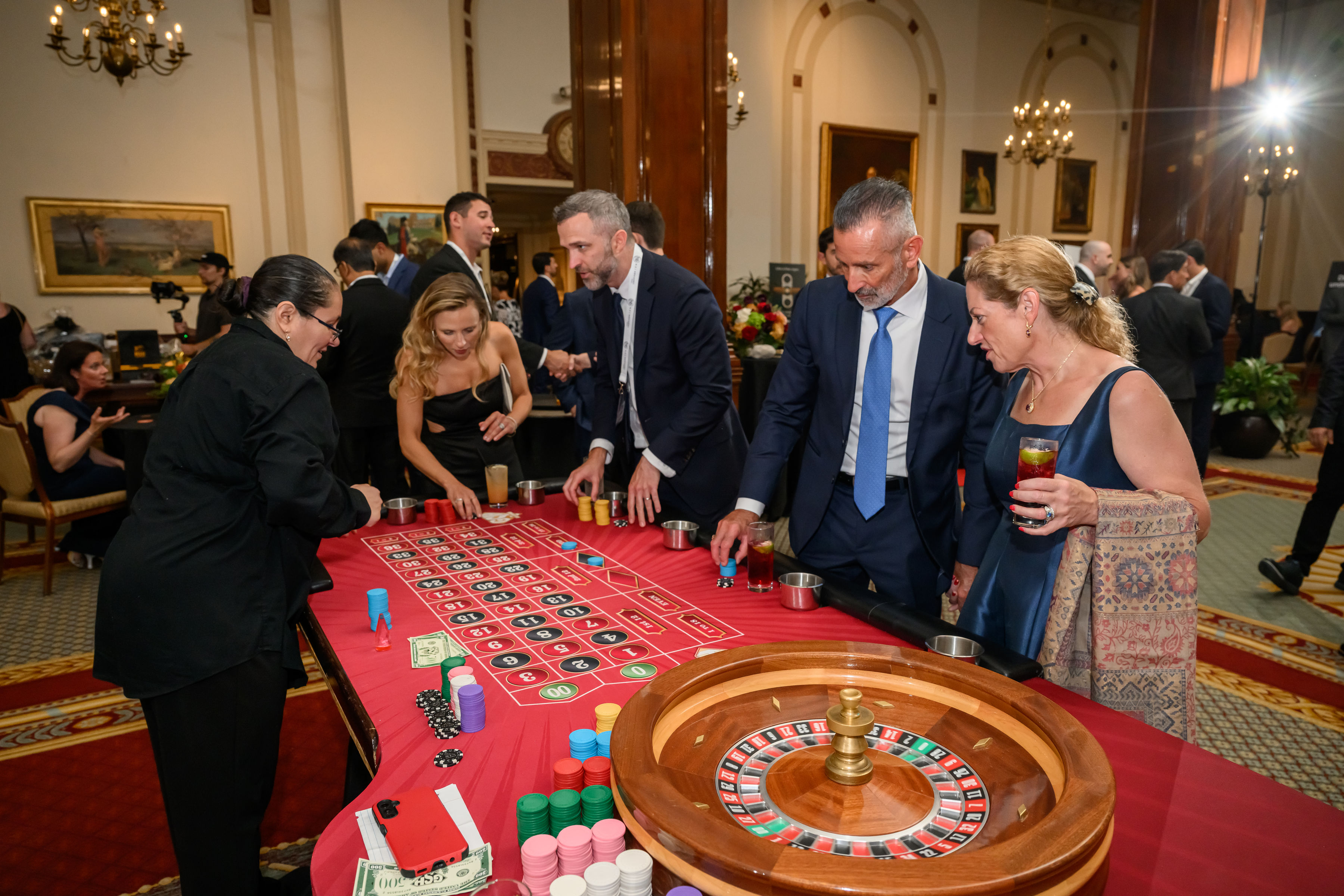 Guests in evening attire gathered around a roulette table at an Astra collective evening reception in a chandelier-lit room.