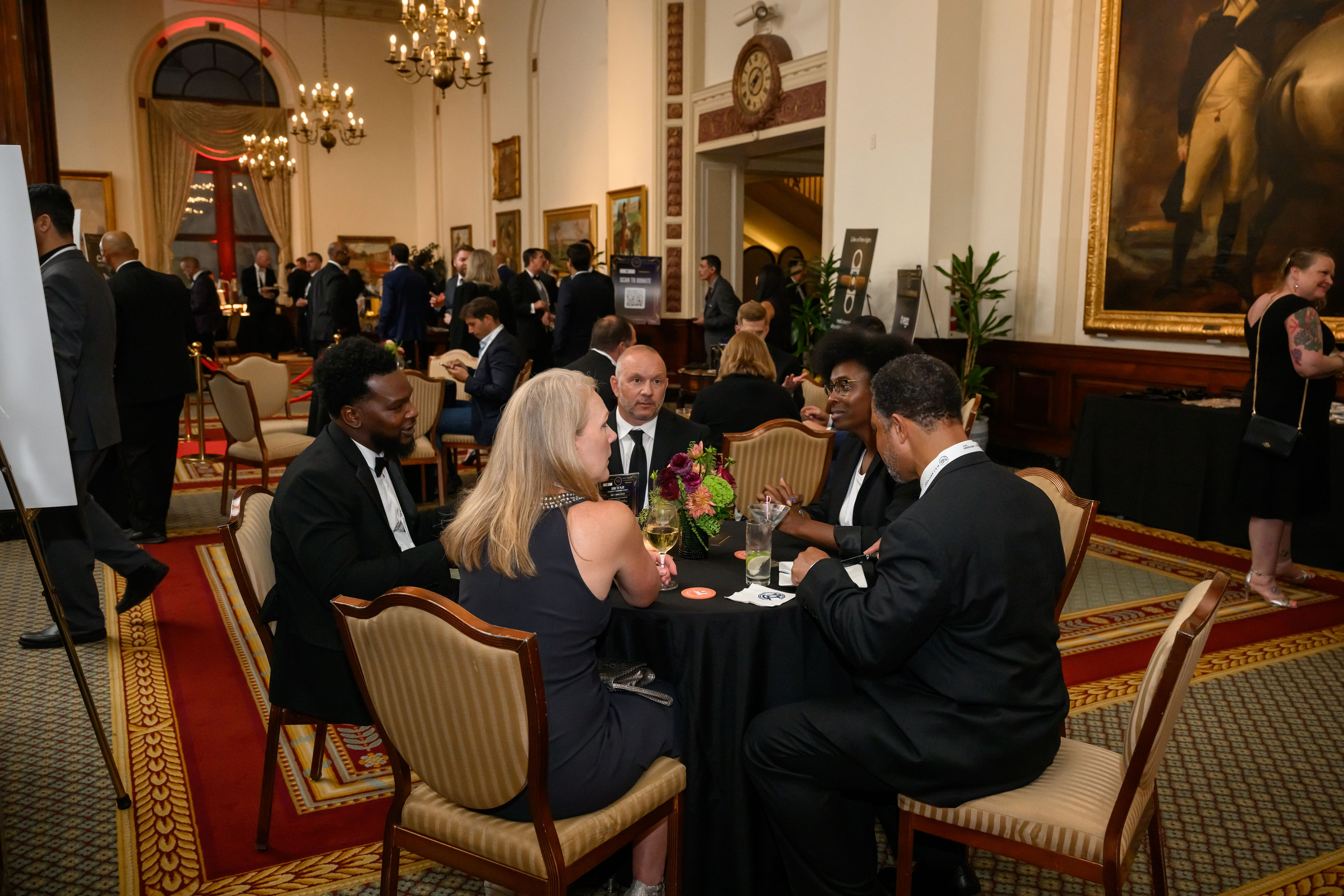 Five guests in black-tie attire in close conversation around a circular table in a chandelier-lit, oak-panelled room.